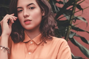 Shoulder-height image of a woman wearing an orange shirt and a charm bracelet, standing in front of large-leafed plants and a red wall